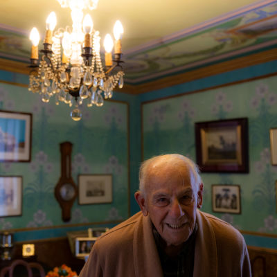 98-vuotias Antonio Grippo kotonaan huoneessa, jossa turkoosit seinät ja kattokruunu sekä seinillä paljon vanhoja valokuvia.