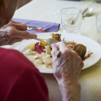 En bild på ett par äldre händer som äter mat med gaffel och kniv.