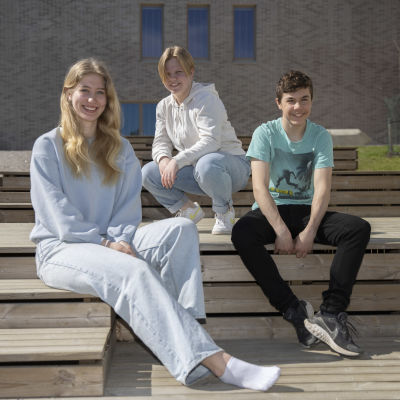 Tre högstadieelever sitter utanför en skolbyggnad i vårsolen och ler mot kameran.