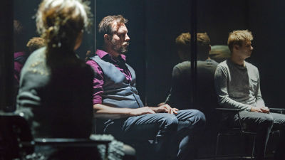Tre skådespelare sitter framför en glasvägg på en mörk scen. 
