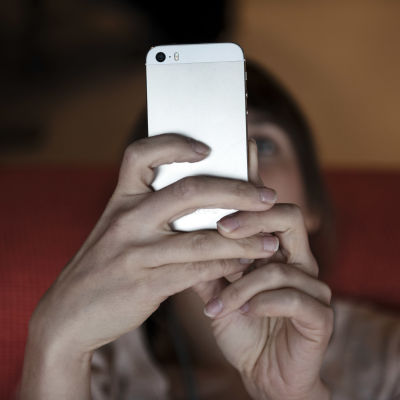 En kvinnas ansikte skymtar bakom en mobiltelefon som hon tittar på. 