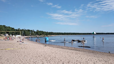 En badstrand en somrig dag. Människor ligger i sanden, badar i havet och åker på sup-brädor i vattnet. I havet skymtar en vattenkarusell.