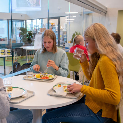Tre flickor sitter vid ett runtbord i Vårberga skolas matsal och äter skollunch. 
