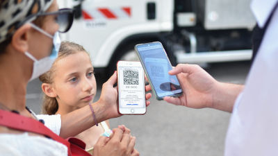 En kvinna med munskydd visar upp sin telefon och sitt digitala coronapass för en kontrollant. I mitten syns en ung flicka som ser på.