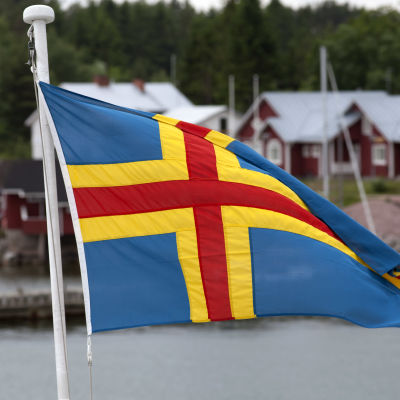 En åländsk flagga i förgrunden, röda hus med vita knutar i bakgrunden.
