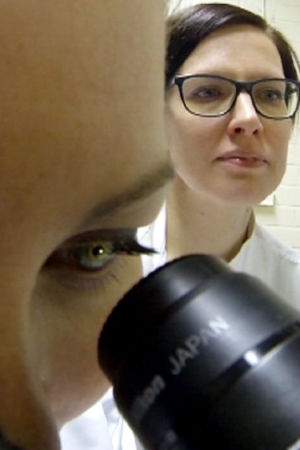 Riina Sarkanen katselee taustalla, kun toinen tutkija katselee mikroskoopilla näytettä.