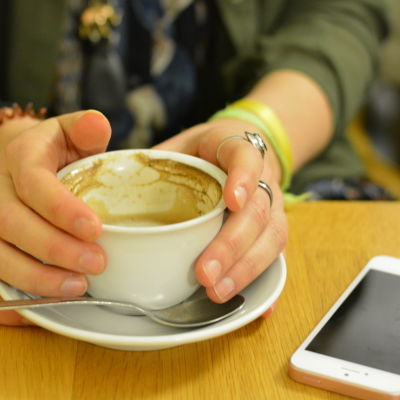 Två händer håller i en kaffekopp med mjölkskum. En telefon ligger bredvid på bordet.