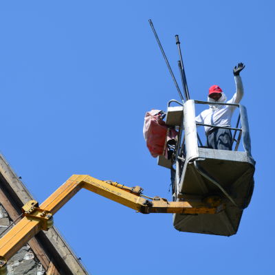 En man iklädd heltäckande kläder står i en skylift bredvid ett tak och vinkar mot kameran.