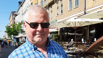 Jack Höglund, en man med kort ljust hår, solglasögon och blårutig skjorta, står på en gågata en solig sommardag.