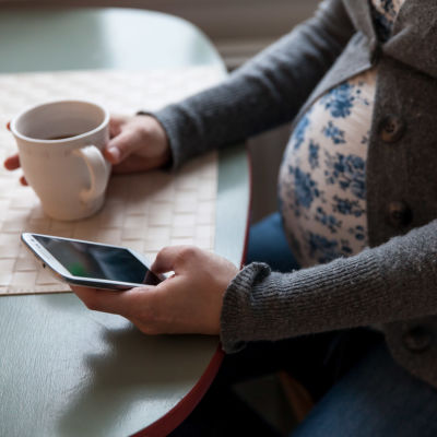 Gravid kvinna sitter och dricker kaffe och använder sin mobiltelefon.