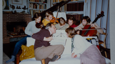 En familj sitter tätt runt en soffa och ett piano. En äldre man och kvinna sitter i soffan och fem barn sitter bakom soffan och spelar varsitt instrument i famnen.