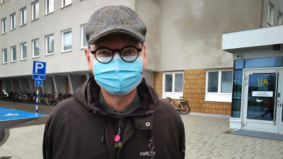 En man med runda svarta glasögon, rutig keps och munskydd står utanför sjukhuset.