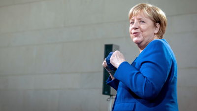 Angela Merkel vänd med sidan mot kameran. Hon tittar förbi kameran och håller i ett munskydd. Hon är klädd i en blå kavaj.