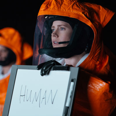 Lingvisten Louise (Amy Adams) är iklädd full skyddsutrustning och håller upp en tavla där hon skrivit "human" / människa och försöker förklara vem hon är för utomjordingarna.