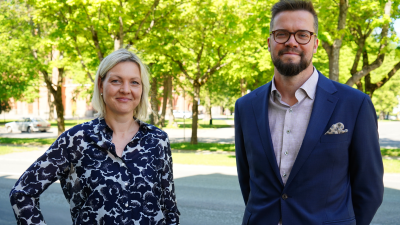 En ljushårig kvinna och en mörkhårig man tittar in i kameran och ler. I bakgrunden syns en solig esplanad.