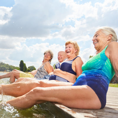 Tre äldre kvinnor och en man sitter på en brygga och plaskar med fötterna i vattnet