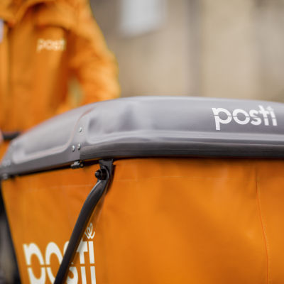 En orange postvagn med texten "Posti" på skuffas av en orangeklädd brevbärare.