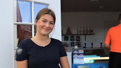 Edit Grönroos, en ung kvinna med brunt hår i hästsvans, ler mot kameran framför en glasskiosk.