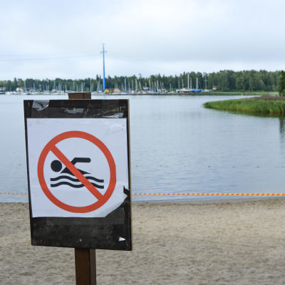 Skylt som visar att det är förbjudet att bada. I bakgrunden syns en avspärrad badstrand.