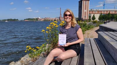 Kvinna sitter på trappa vid havet och håller i en bok. Hon ler mot kameran.