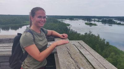 Kvinna står uppe i utkikstorn. Hon ler stort. I bakgrunden är hav och skog