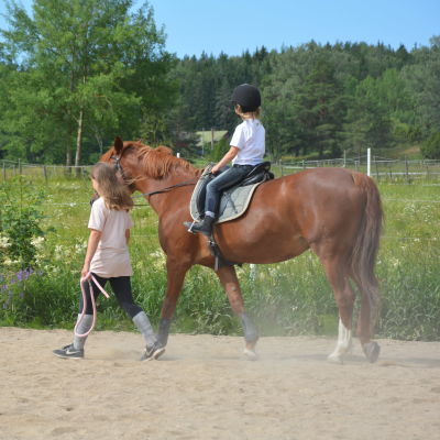 En tolvårig flicka leder en häst som en mindre flicka rider på, på en sandplan.