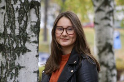Kvinna i glasögon ler mot kameran med björkar i bakgrunden.