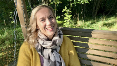 Leende kvinna med blont lockigt hår och gul kappa sitter i trädgårdsgunga.