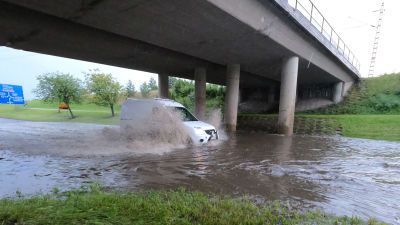 En vit minivan kör genom djupt flödvatten under en bro.