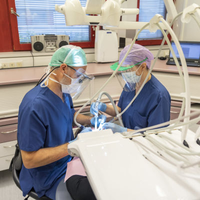 Två tandläkare undersöker en patients tänder.Tandläkarna har munskydd och visir framför ansiktet.