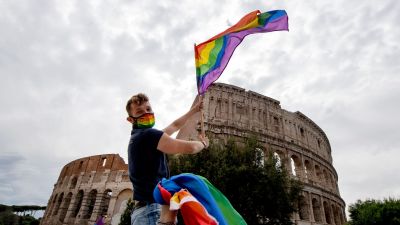 En man sitter på ett biltak och håller i en prideflagga. I bakgrunden romska colosseum.