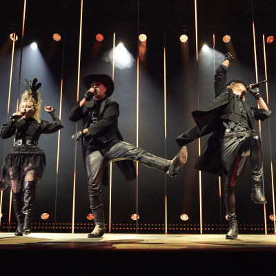 Fyra sångare klädda i svarta scenkläder dansar på en dramatiskt upplyst scen. 