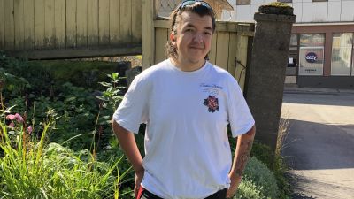En ung man med vit t-skjorta står på en gård med blommor. Bakom ett hus med skyltfönster där det står "Eezy vapaus työhön".