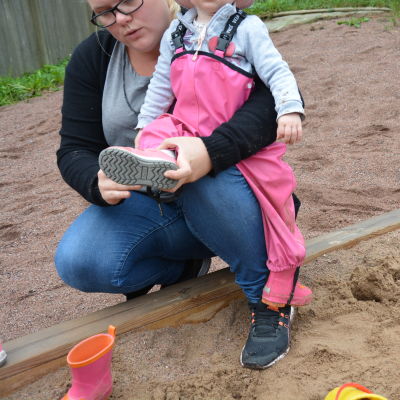 En dagistant klär på stövlar på ett barn i sandlådan.