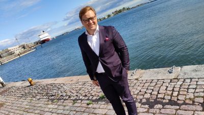 En ung man med ljust hår står i kostym framför havet. I bakgrunden syns två fartyg. Han ser in i kameran och ler.