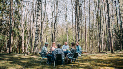 Kuvan etualalla kuusi henkilöä istuu pöydän ääressä ulkosalla kahvilla. Taustalla keväinen koivikko ilman lehtiä.