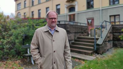En man i beige jacka står framför ett gult stenhus.