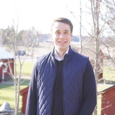 en ung man men börk hår som ler mot kameran, i bakgrunden syns en åker och några röda hus.