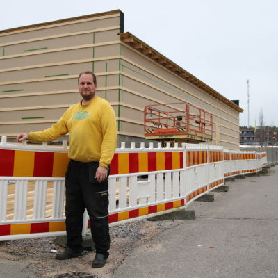 En man i gul tröja står utomhus vid ett bygge. Röd-gult staket runt bygget.