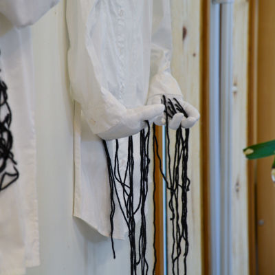 En vit kragskjorta hänger på en vägg. Vid ärmarna finns två händer gjorda av textil som det hänger svarta trådar ifrån.