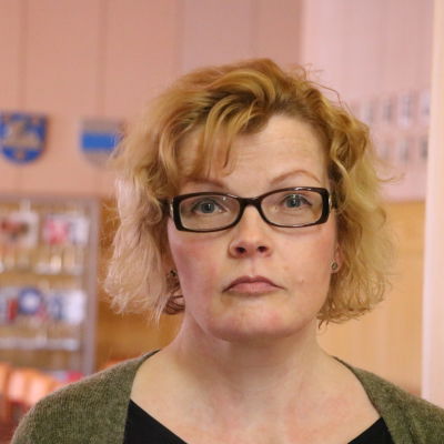 En kvinna med axellångt ljus hår och glasögon står i en dörröppning. 