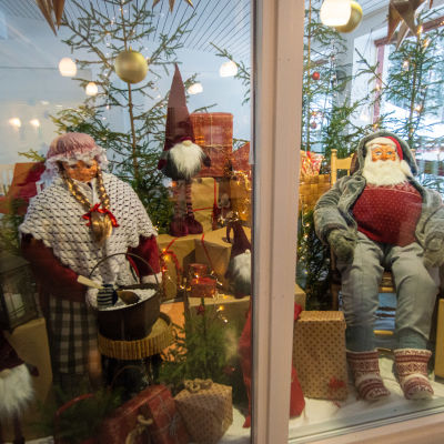 Två stora dockor föreställande julgubben och julgumman sitter omgivna av julklappar.