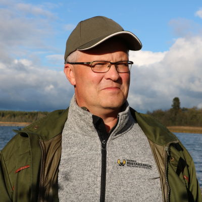 En medelålders man med keps och glasögon står framför havet. Han är iklädd en grön jacka och grå fleece-tröja med texten Finlands viltcentral skrivet på vänstra bröstet.