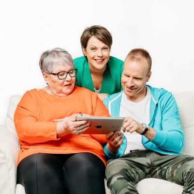 Oranssissa paidassa oleva nainen ja Mikko Kekäläinen istuvat sohvalla ja katsovat tablettia. Anna-Liisa Tilus seisoo takana, katsoo kameraan ja hymyilee.
