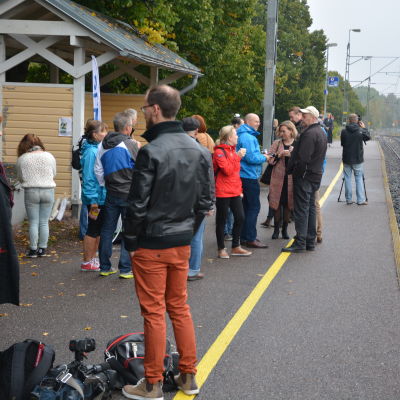 Människor står på en tågperrong, tågbana till höger, höstlöv på marken.