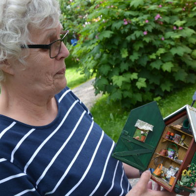 elisabeth westerholm står i trädgården och håller ett tittskåp i händerna. Skåpet är en miniatyrmodell av ett redskapsskåp för en trädgård. 