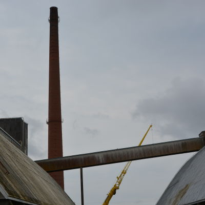 På bilden ser man skorstenen på avstånd mellan två sädessilon. Bredvid skorstenen står en gul lyftkran. 