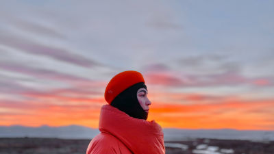 Meeri Koutaniemi katsoo kuvan oikean reunaan. Hän seisoo lähellä kameraa punainen paksu takki ja punainen hattu päässään. Taivaalla auringonnousu.