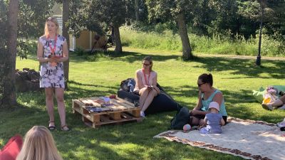 En kvinna i klänning står och pratar framför kvinnor som sitter i gräset på picnic