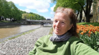 Marjan Raar, en dam med bruntgrått bakåtkammat hår, scarf och en grön tröja, sitter på en bänk vid åstranden med tulpaner bakom sig.